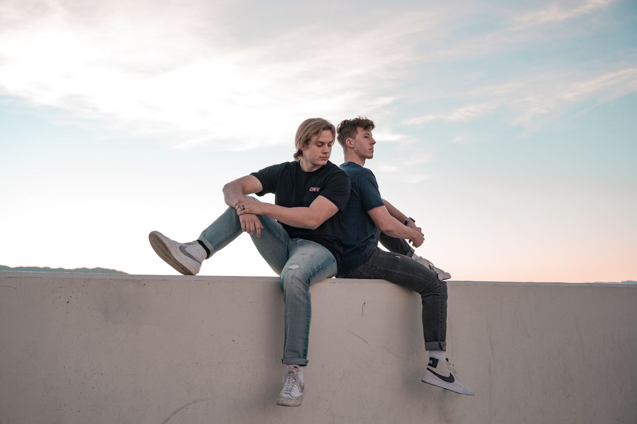 Services Two young men sitting back to back on a wall under a clear evening sky.