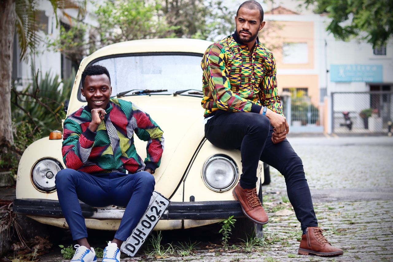 Services Two men wearing vibrant African print shirts posed with a classic yellow Volkswagen Beetle outdoors.