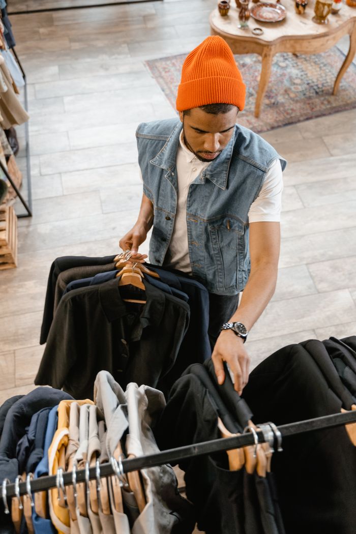 About Man examines clothing in boutique wearing denim vest and orange beanie.